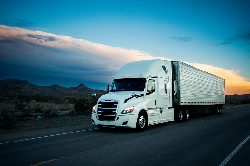 White semi-truck traverses a 2-lane highway in the Arizona desert at dusk, its imposing frame set against a dramatic cloudscape, as it journeys through the rugged terrain White semi-truck traverses a 2-lane highway in the Arizona desert at dusk, its imposing frame set against a dramatic cloudscape, as it journeys through the rugged terrain